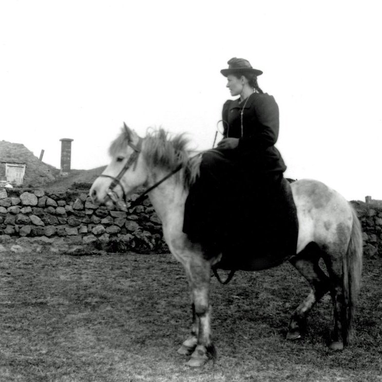 Kona á hestbaki við torfbæ. Óþekkt. Myndin tekin ca. 1898-1912.