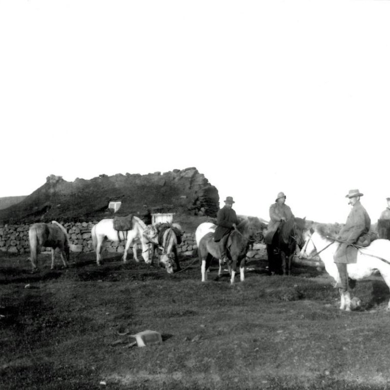 Hópur fólks á hestum. Útihús í bakgrunni.  Óþekkt. Myndin tekin ca. 1898-1912.