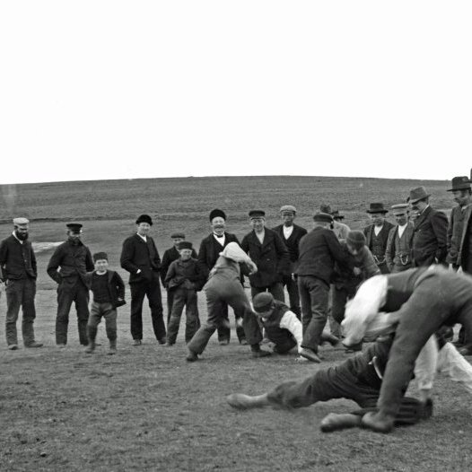Glímt úti á Höfða. Glíma var allmjög iðkuð á Husavík í byrjun aldarinnar og sjadnast skorti áhorfendur. Nokkrir iðkuðu grísk-rómverska glímu. Líkur eru á að þessi mynd sé tekin 14. júní 1905 en þá var haldinn skemmtifundur úti á Höfða.